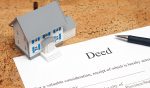 A miniature house sits on a table along with a paper deed and a pen.